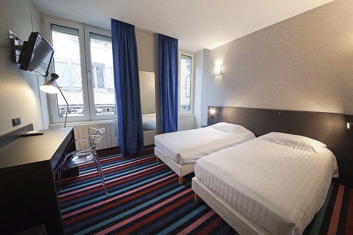 Ett hotellrum med två sängar, ett skrivbord, en stol, en tv som är monterad på väggen och ett fönster med gardiner.