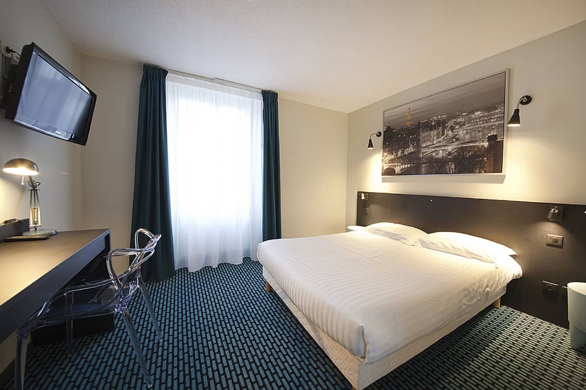 Ett hotellrum med en säng, ett skrivbord, en stol, en tv och en inramad tavla på väggen.