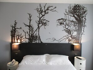 Habitación de hotel con cama, ropa de cama blanca, un cuadro colgado en la pared con árboles y dos lámparas de noche.