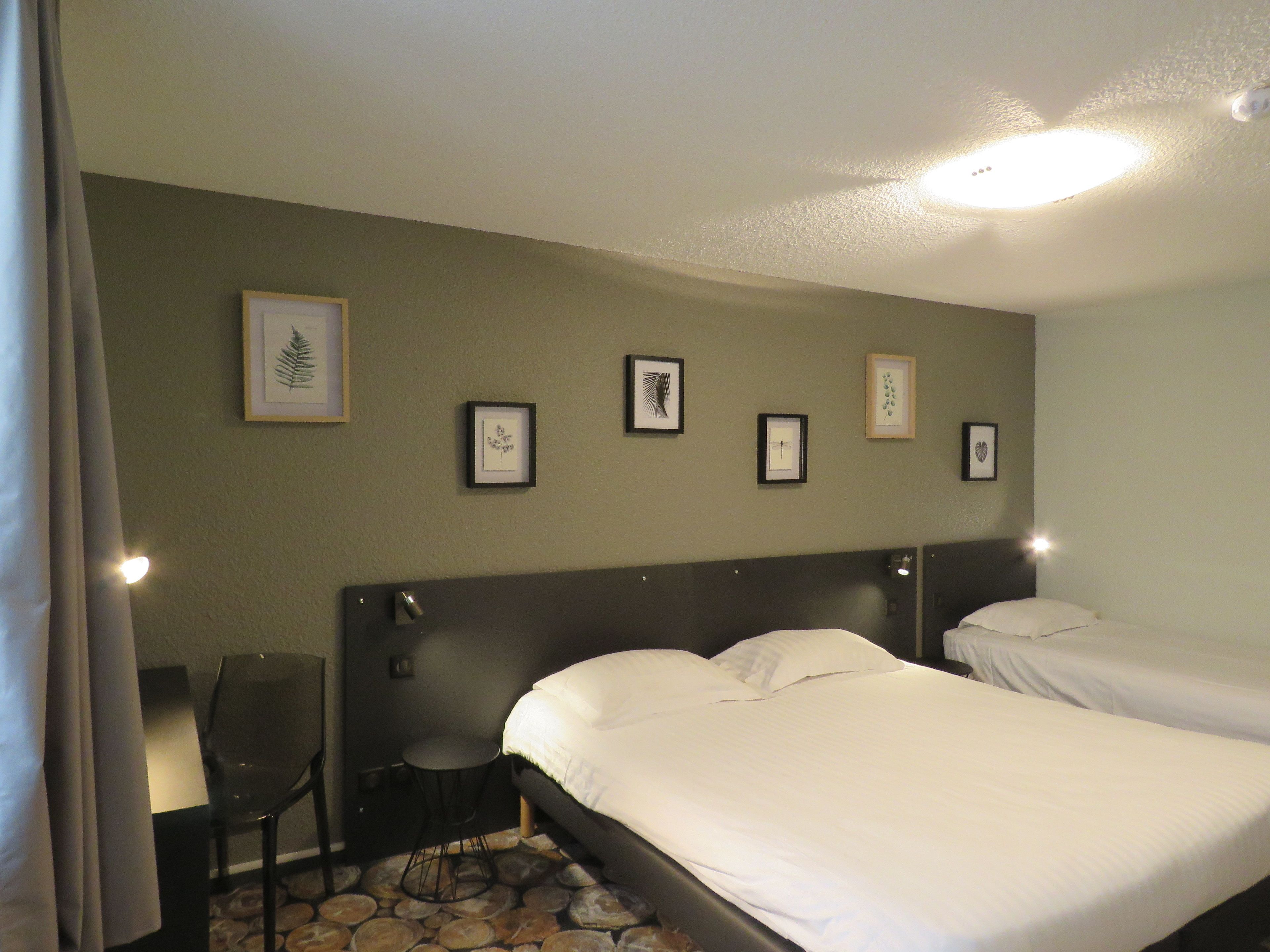 A hotel room with two beds, a small table, a chair, and framed pictures on the wall.