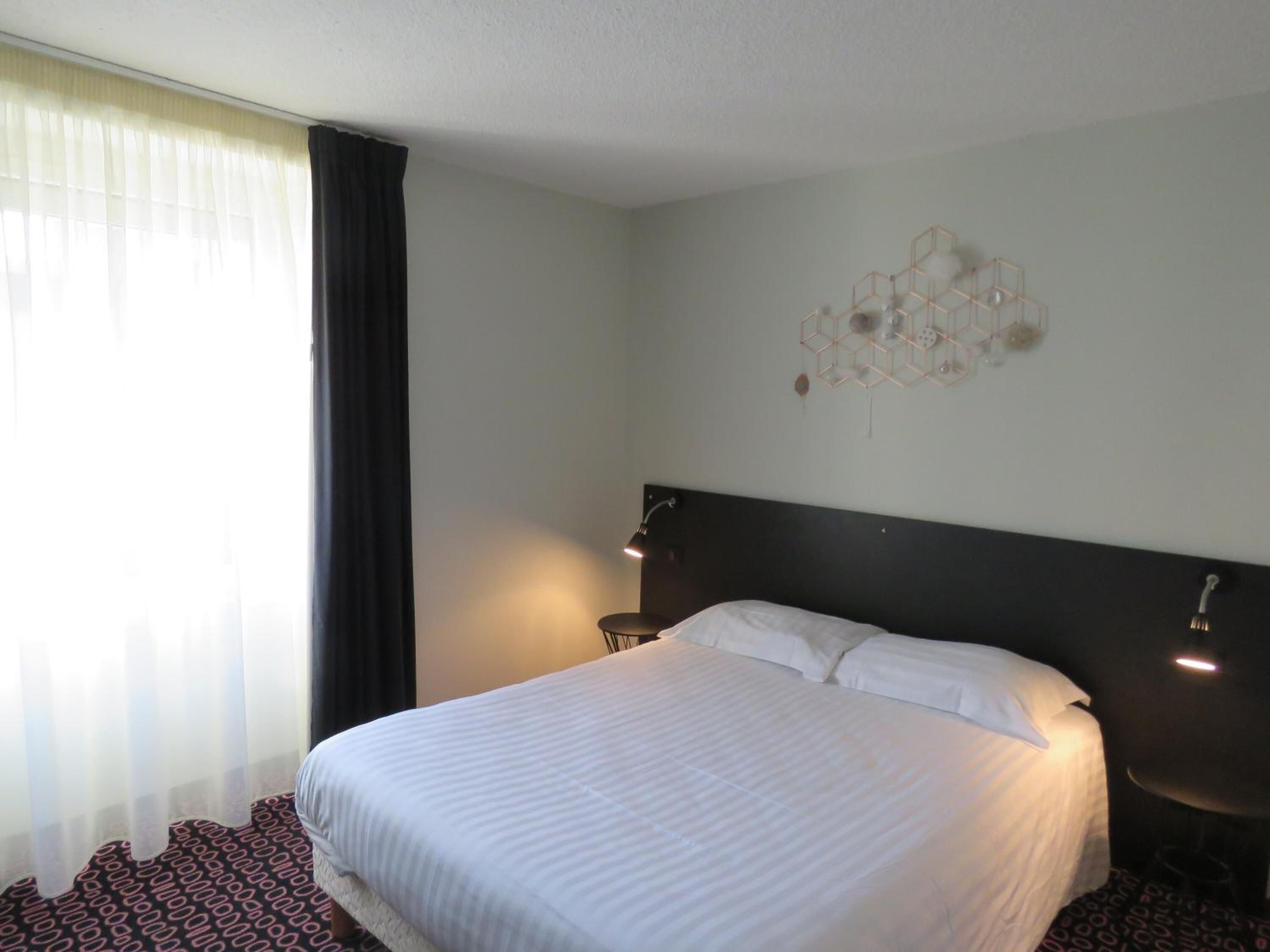 Ein Hotelzimmer mit einem großen Bett, einem dunklen Kopfteil, einer Wandleuchte und einem Fenster mit transparenten Vorhängen.