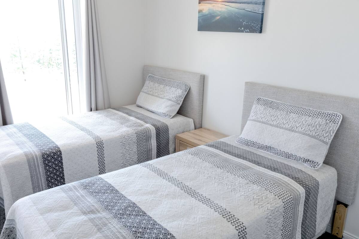 Una habitación con dos camas, cada una con cabecero gris y colcha estampada. Hay una mesita de noche de madera entre las camas.