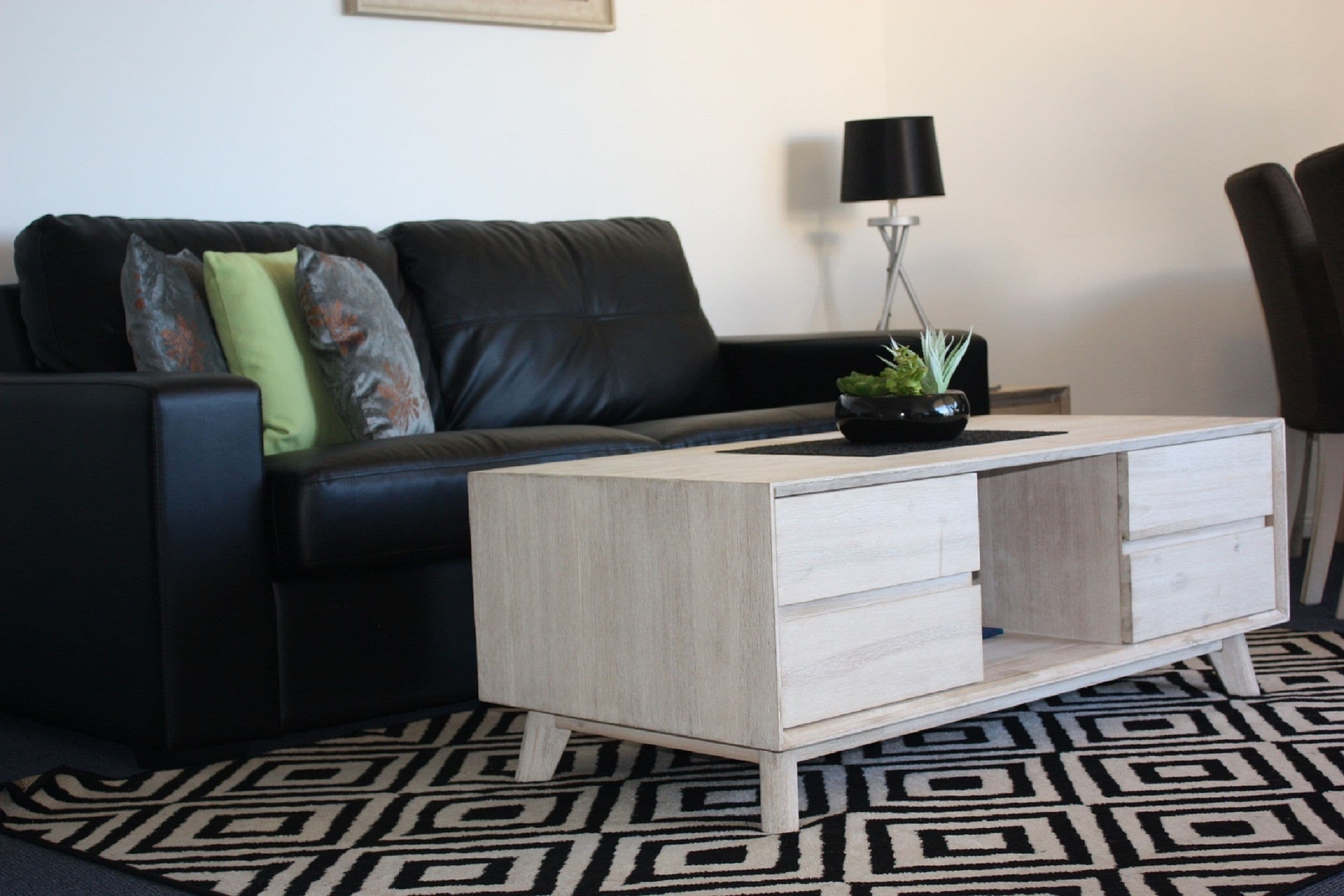 Un salon moderne avec un canapé en cuir noir, une table basse en bois clair dotée de tiroirs, un lampadaire noir et un tapis à motifs.