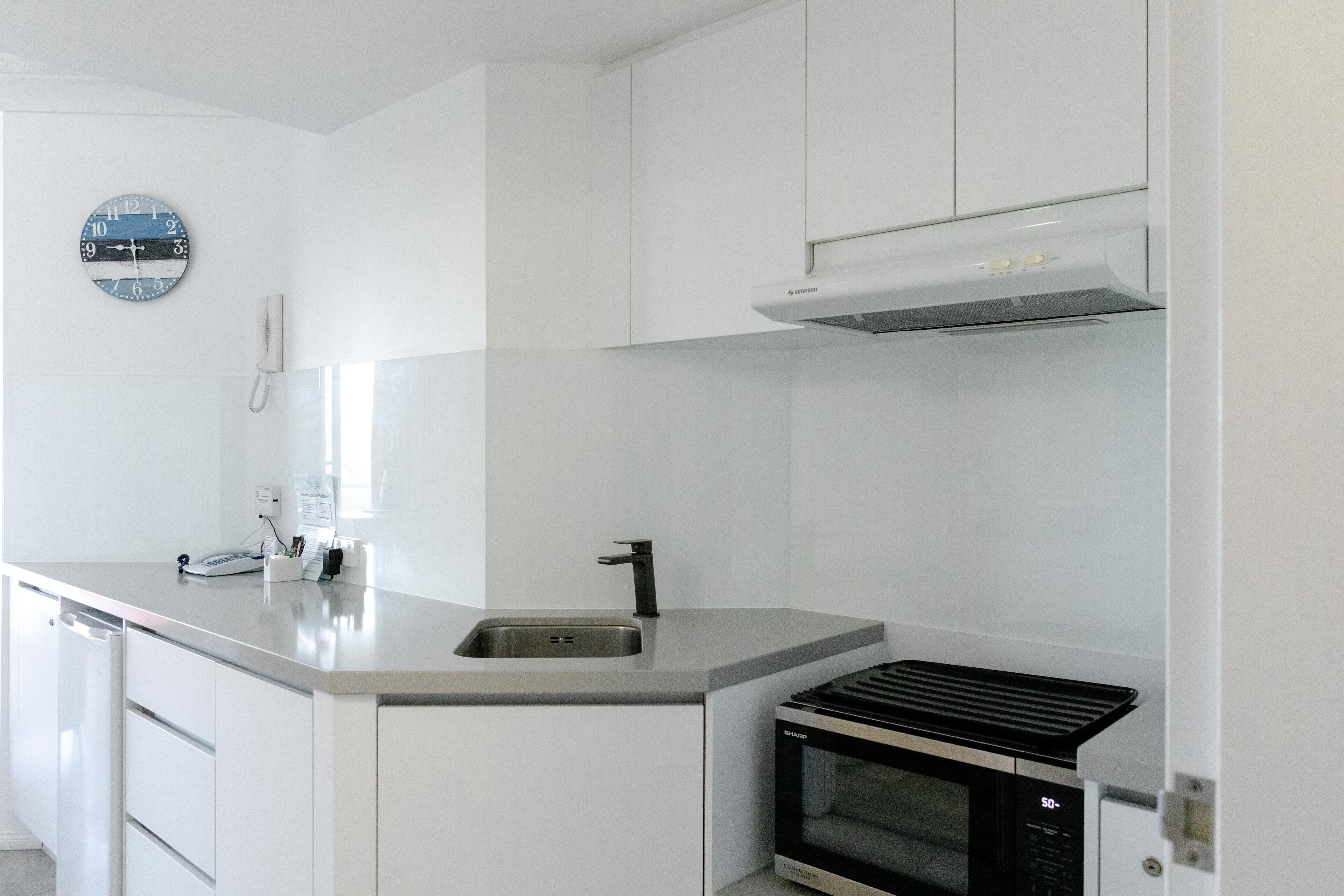 A modern kitchen with white cabinetry, a stainless steel sink, a black microwave, and a clock on the wall.