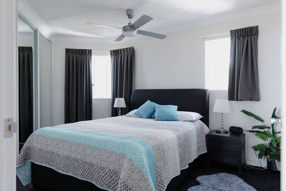 Un dormitorio con una cama, una mesita de noche, una lámpara, un ventilador de techo y una ventana con cortinas.