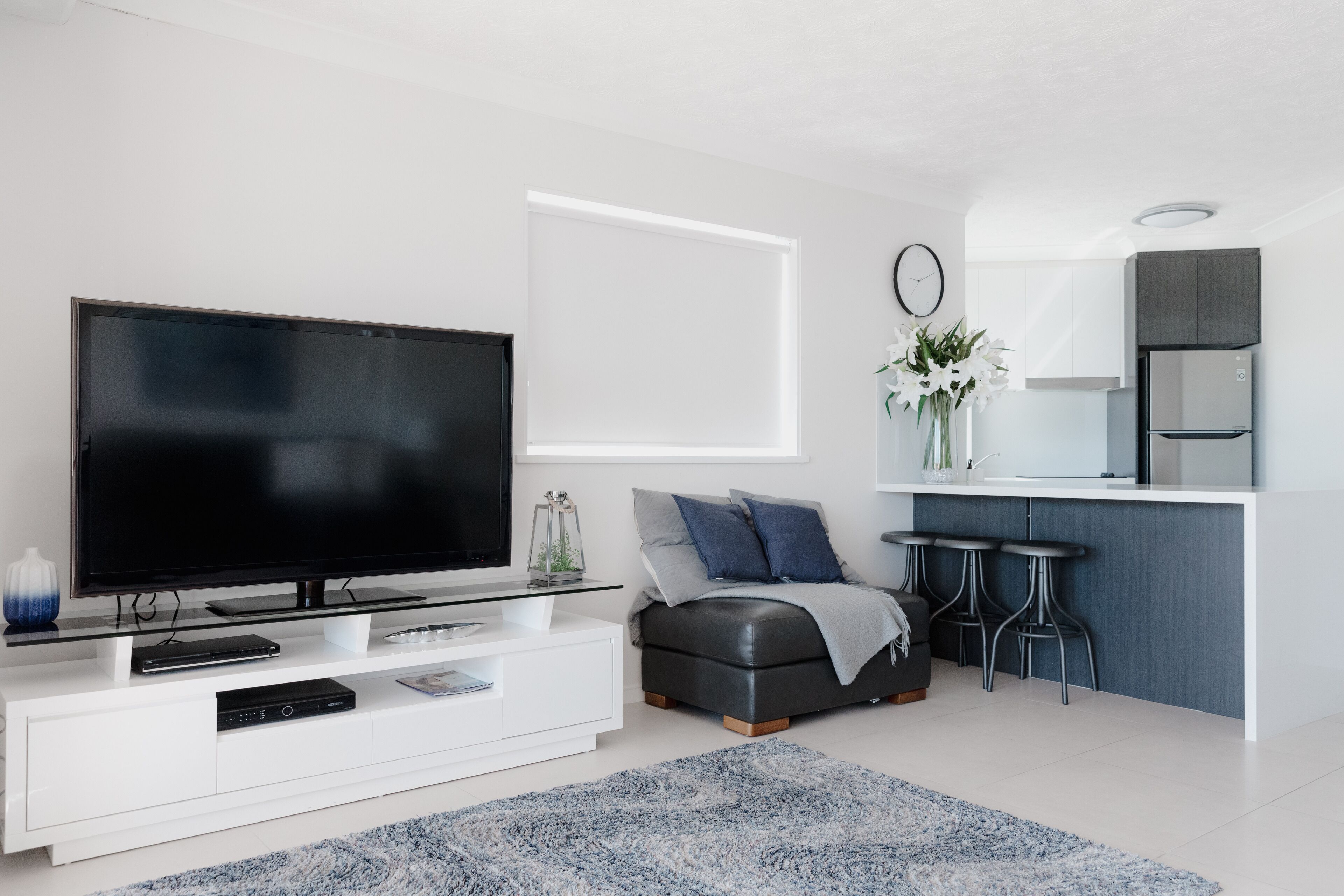 A modern living room with a large flat-screen TV, a glass coffee table, a sofa with cushions, a small ottoman, and a kitchen area with a fridge and bar stools.