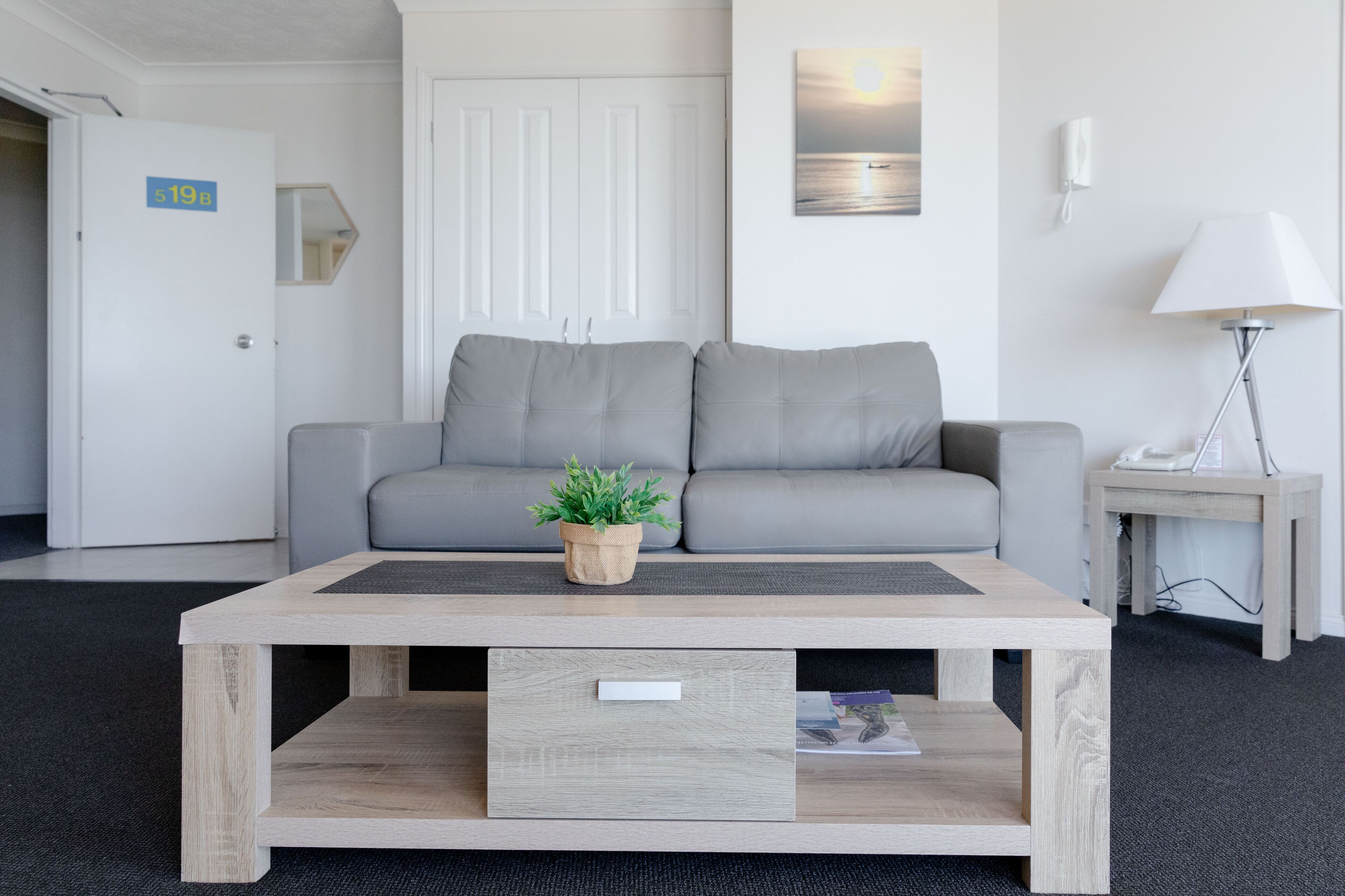 Une pièce moderne avec un canapé gris, une table basse en bois dotée d’un tiroir, une petite plante en pot, une lampe fixée au mur et un tableau encadré.
