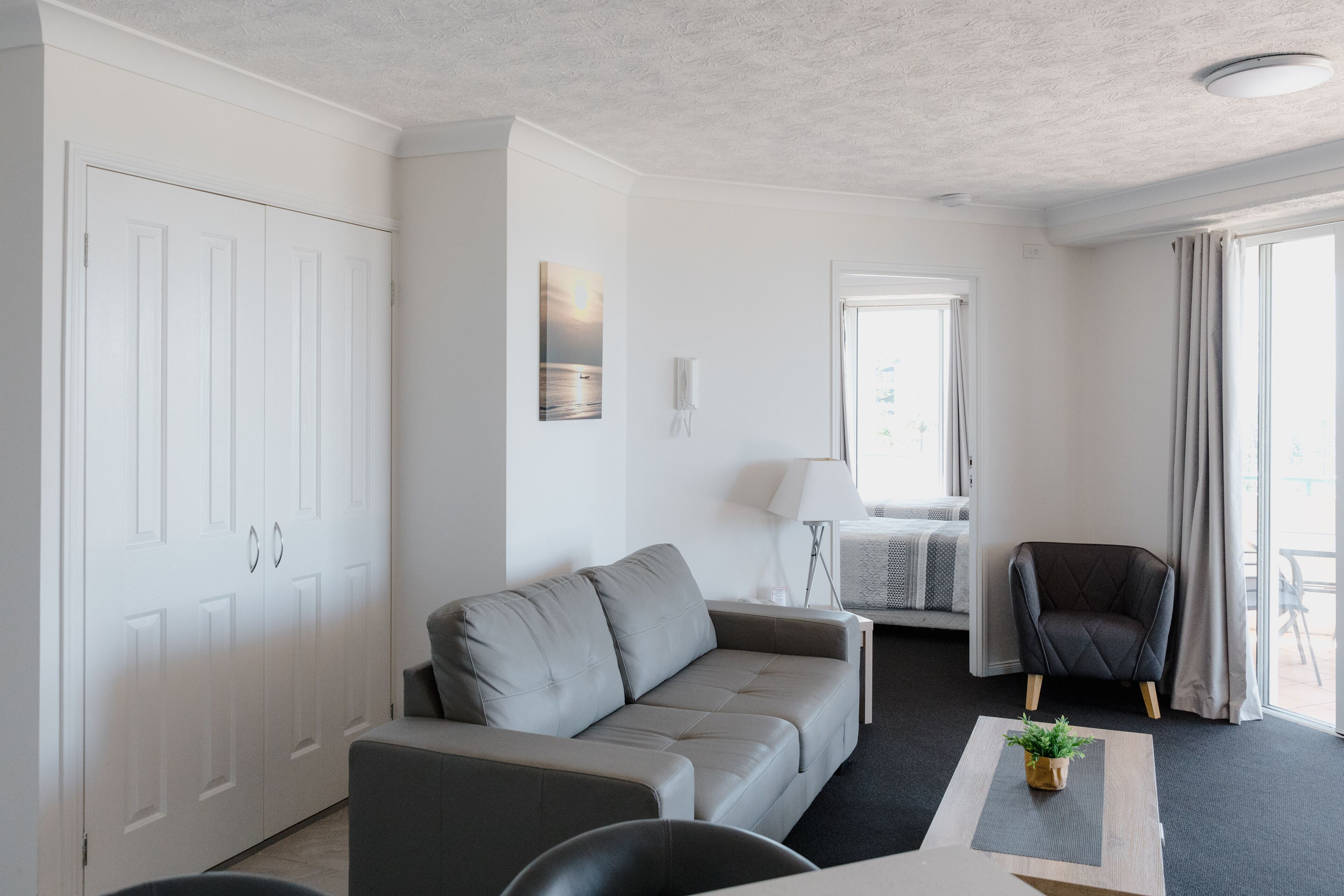 A modern, minimalist living room with a grey sofa, a small table with a plant, a dark armchair, and a white wardrobe.