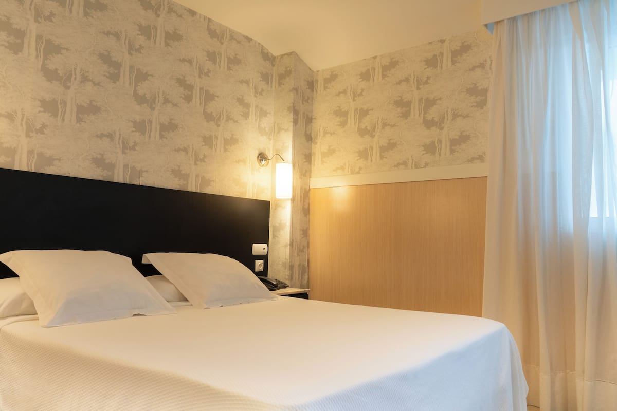 A hotel room with a bed, a nightstand, a wall-mounted lamp, and a window with curtains.