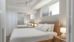 Quarto com cama de madeira, roupa de cama branca, mesa de cabeceira com abajur, espelho e ventilador de teto.