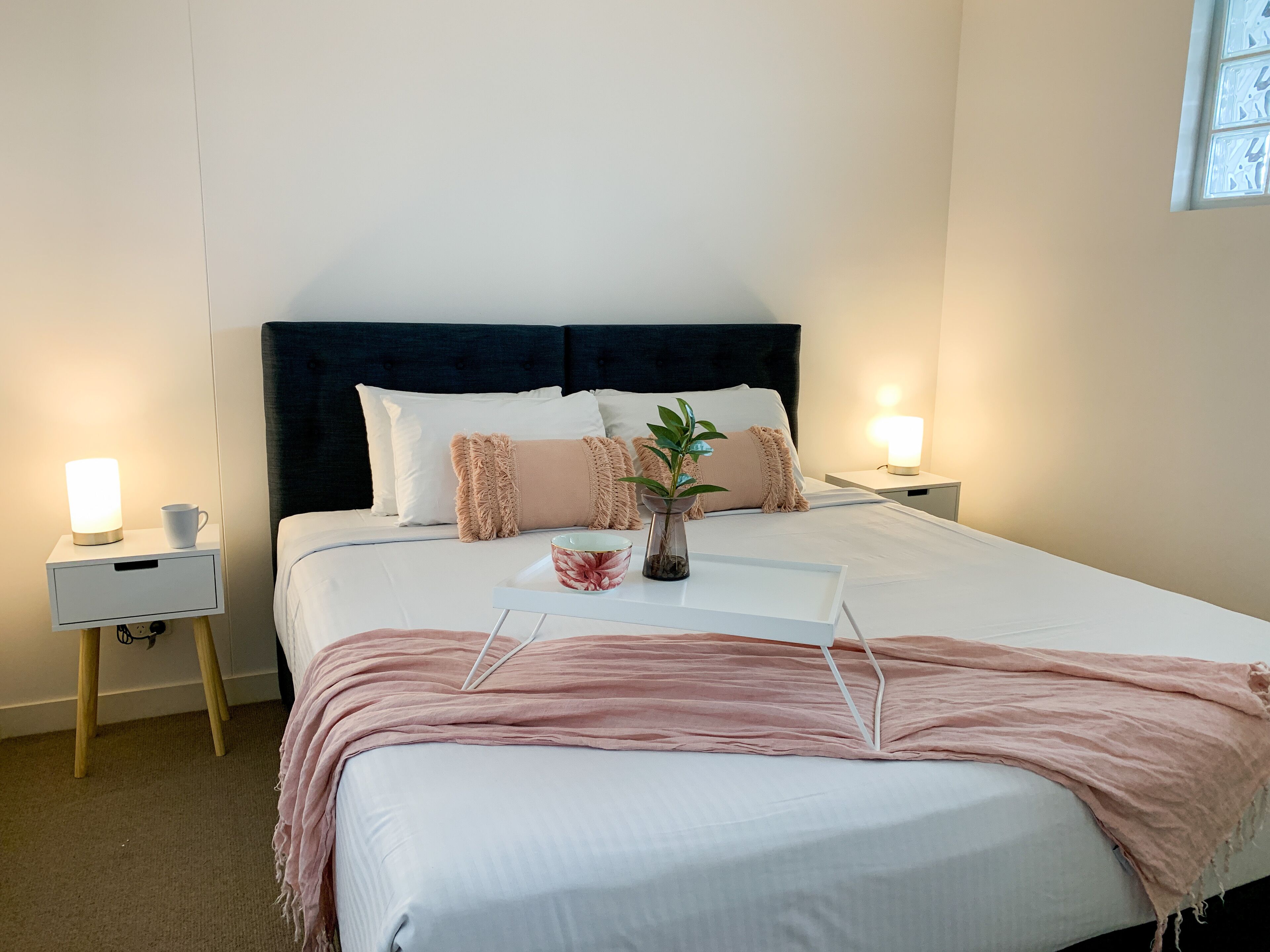A neatly made bed with a dark headboard, two bedside tables with lamps, a bedside plant, and a pink throw blanket.