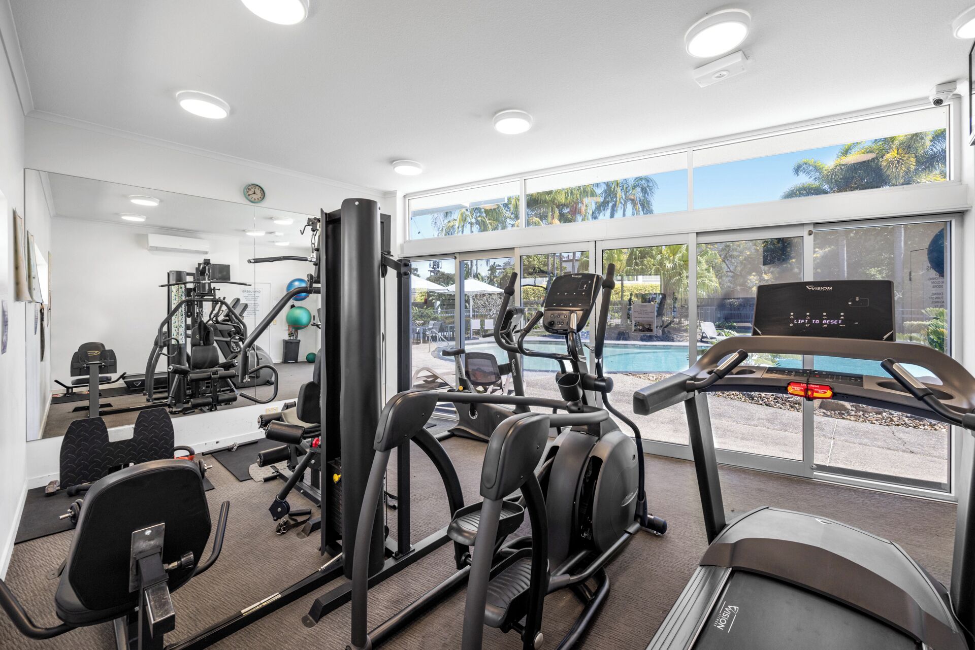 A gym with various exercise equipment, a large window with a view of a swimming pool and palm trees, and a clock on the wall.