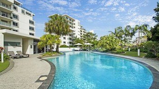 Piscina cercada por área ajardinada com palmeiras e um edifício alto ao fundo.