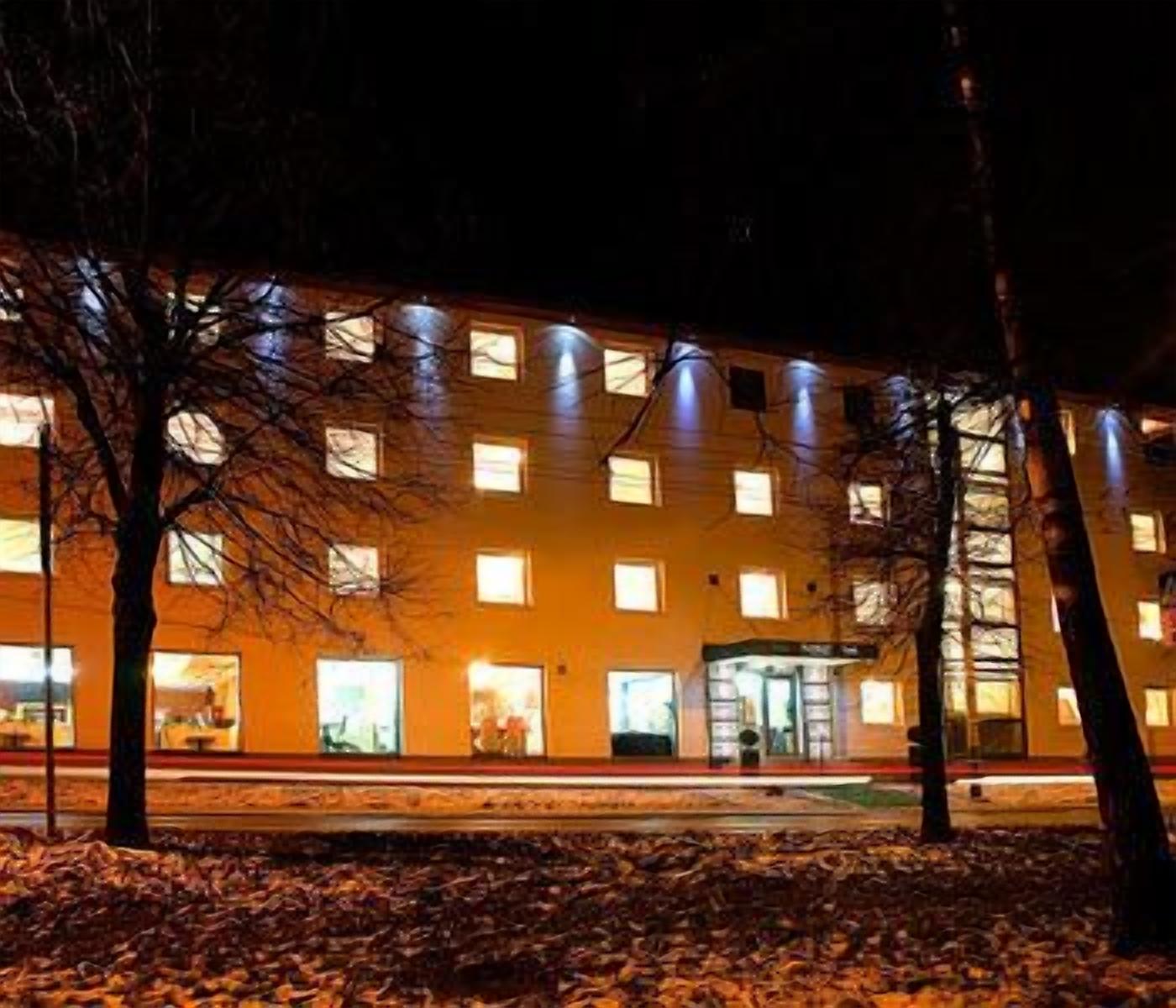 A multi-story building with illuminated windows at night, a visible entrance, and a parking area in front.