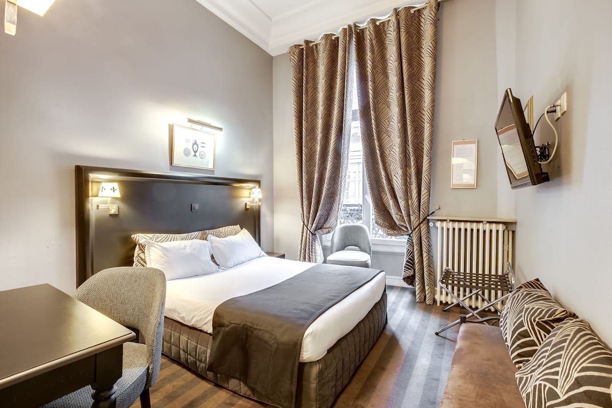 Habitación de hotel con cama, escritorio, silla, sofá, radiador y cortinas.