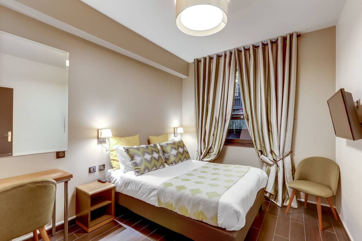 Una habitación de hotel con cama, mesitas de noche, una silla, un escritorio, un espejo y cortinas.
