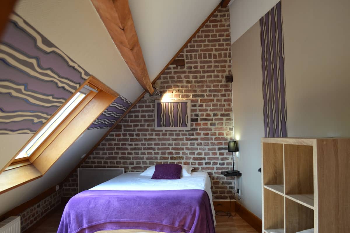 A bedroom with a sloped ceiling, exposed brick walls, a bed with a purple blanket, a wooden wardrobe, and a wall-mounted light fixture.
