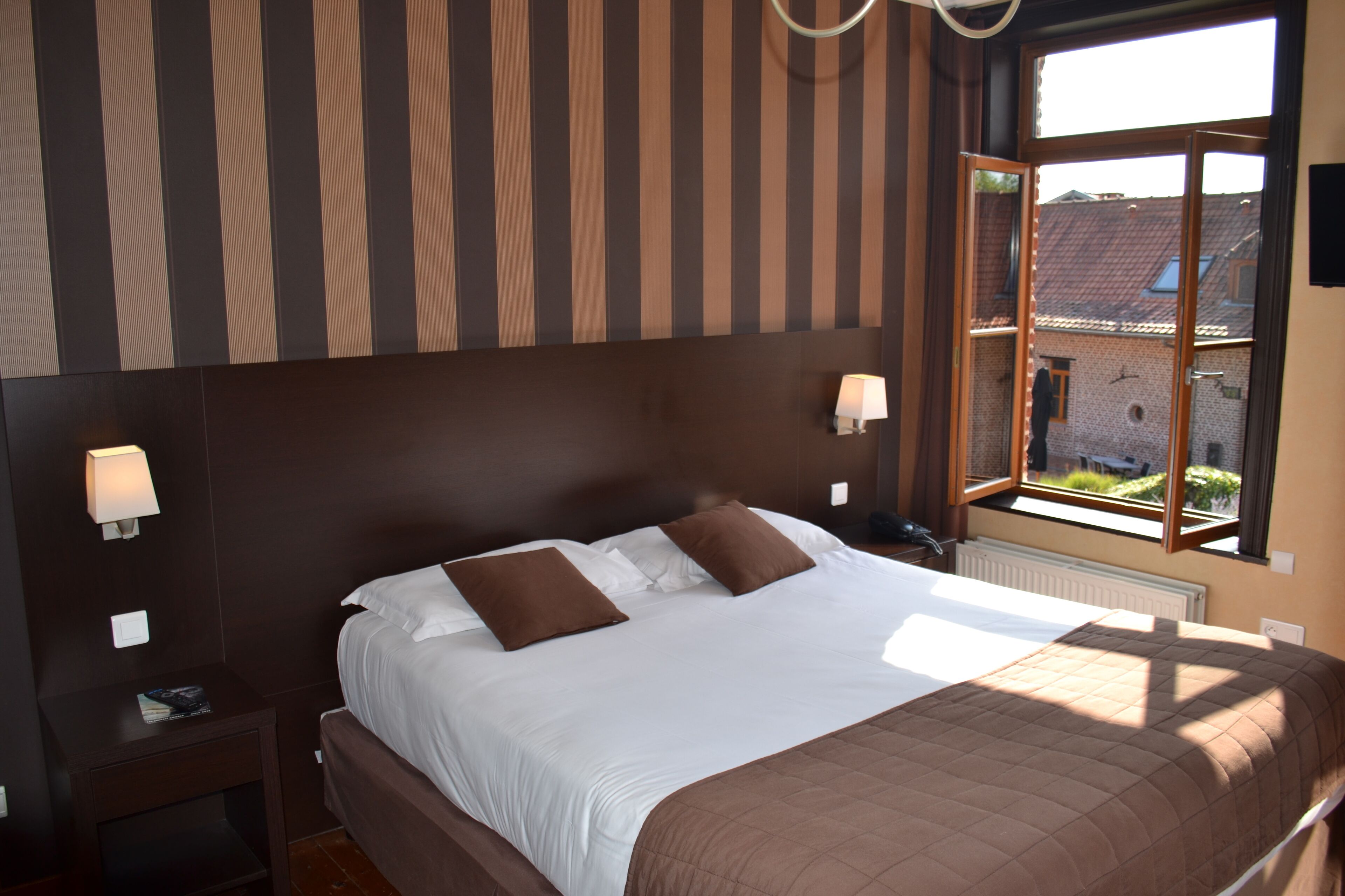 A bedroom with a large bed, two bedside lamps, a window with a view of buildings, and a striped wall.