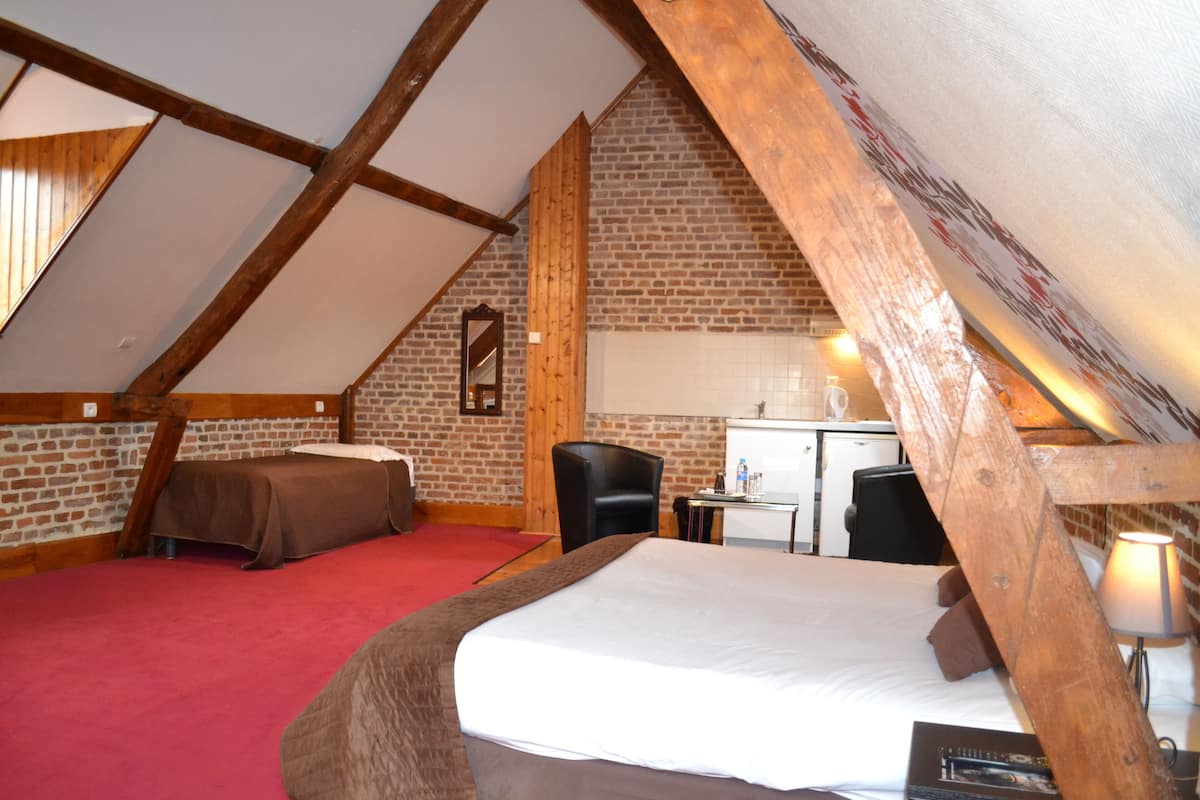 A room with a sloped ceiling, exposed wooden beams, a brick wall, a bed with a red carpet, a desk, and a chair.