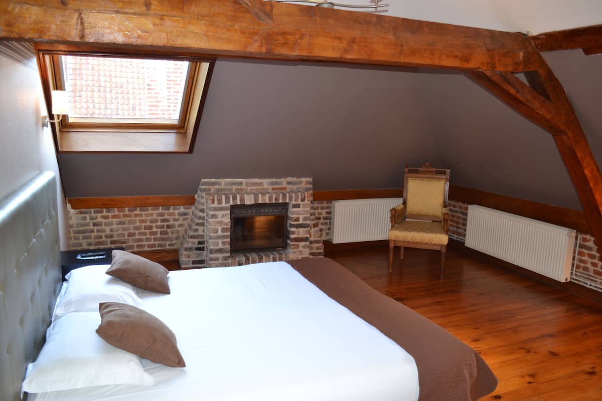 A bedroom with a brick fireplace, a bed with pillows, a chair, and wooden beams.
