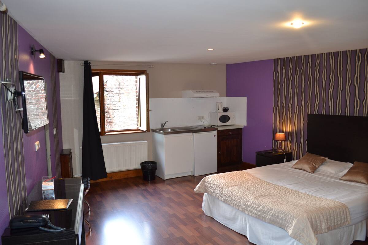 A bedroom with a bed, a nightstand, a desk, a window, and a kitchenette.