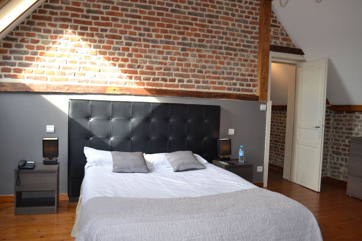 A bedroom with a brick wall, a tufted headboard, a wooden floor, a bedside table with a lamp, and a nightstand with a phone.