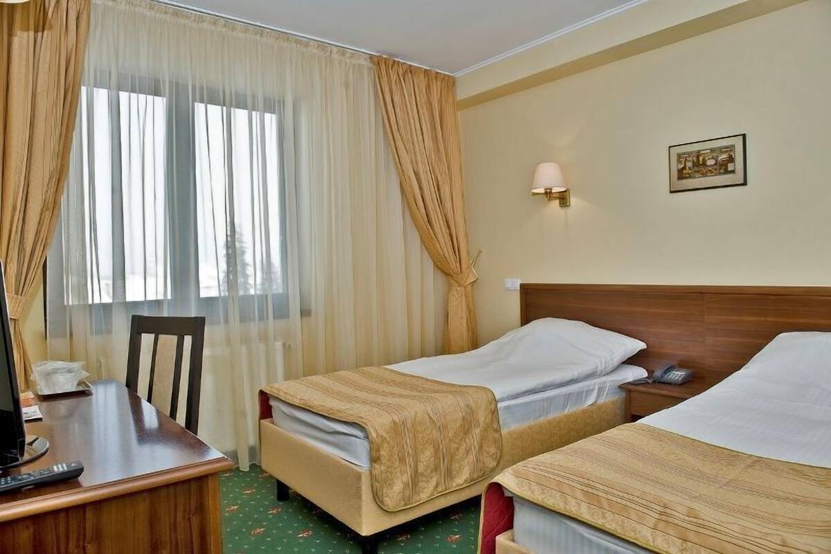 Habitación de hotel con dos camas individuales, un escritorio con silla, una mesita de noche con lámpara y una ventana con cortinas translúcidas.
