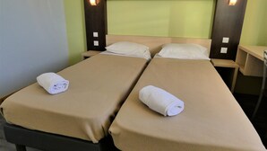 A double room with two beds, each with a beige bedspread and a rolled white towel. There are bedside tables with lamps and wall-mounted light fixtures.