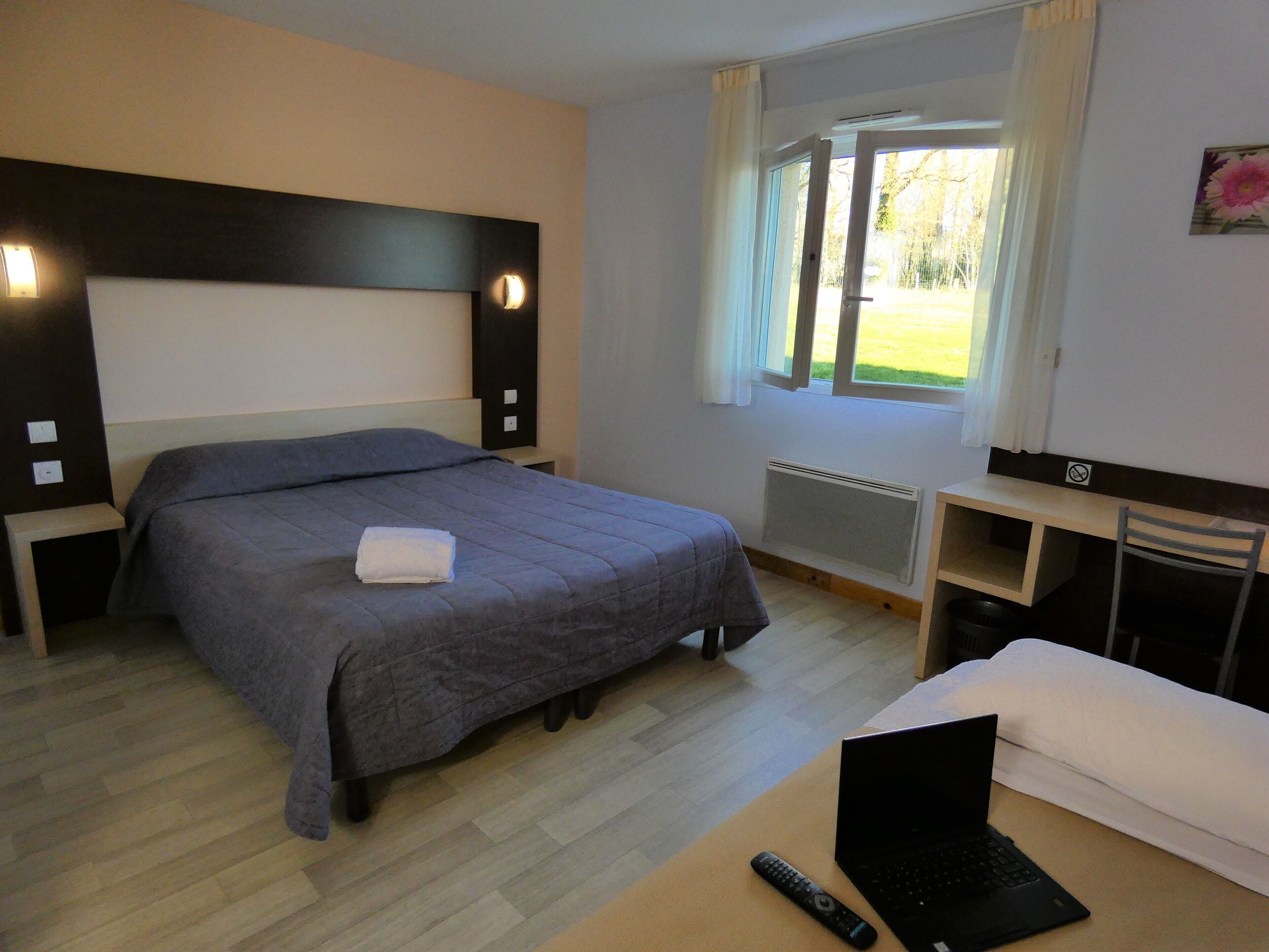 A hotel room with a bed, a desk with a laptop, a remote control, and a window with curtains.