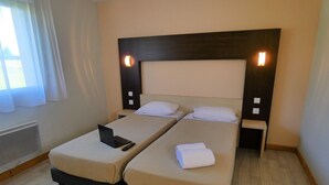 A hotel room with two single beds, a wooden headboard, bedside lamps, a laptop on one bed, and a folded towel on the other.