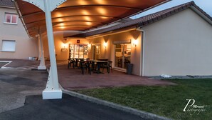 A covered outdoor seating area with tables and chairs, a trash bin, and a building entrance.