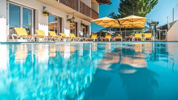 Une piscine avec des chaises longues jaunes et des parasols, un balcon avec une balustrade, et un grand arbre en arrière-plan.