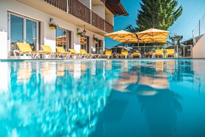 A swimming pool with yellow lounge chairs and umbrellas, a balcony with a railing, and a large tree in the background. - Le Christania (Bellevaux)