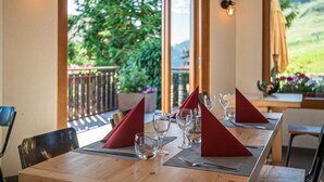 Une table à manger dressée avec des serviettes rouges, des verres transparents et des couverts, donnant sur un paysage verdoyant et une terrasse en bois à l’extérieur.