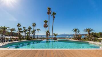 Un área de piscina con cubierta de madera, rodeada de palmeras y un cielo azul despejado.