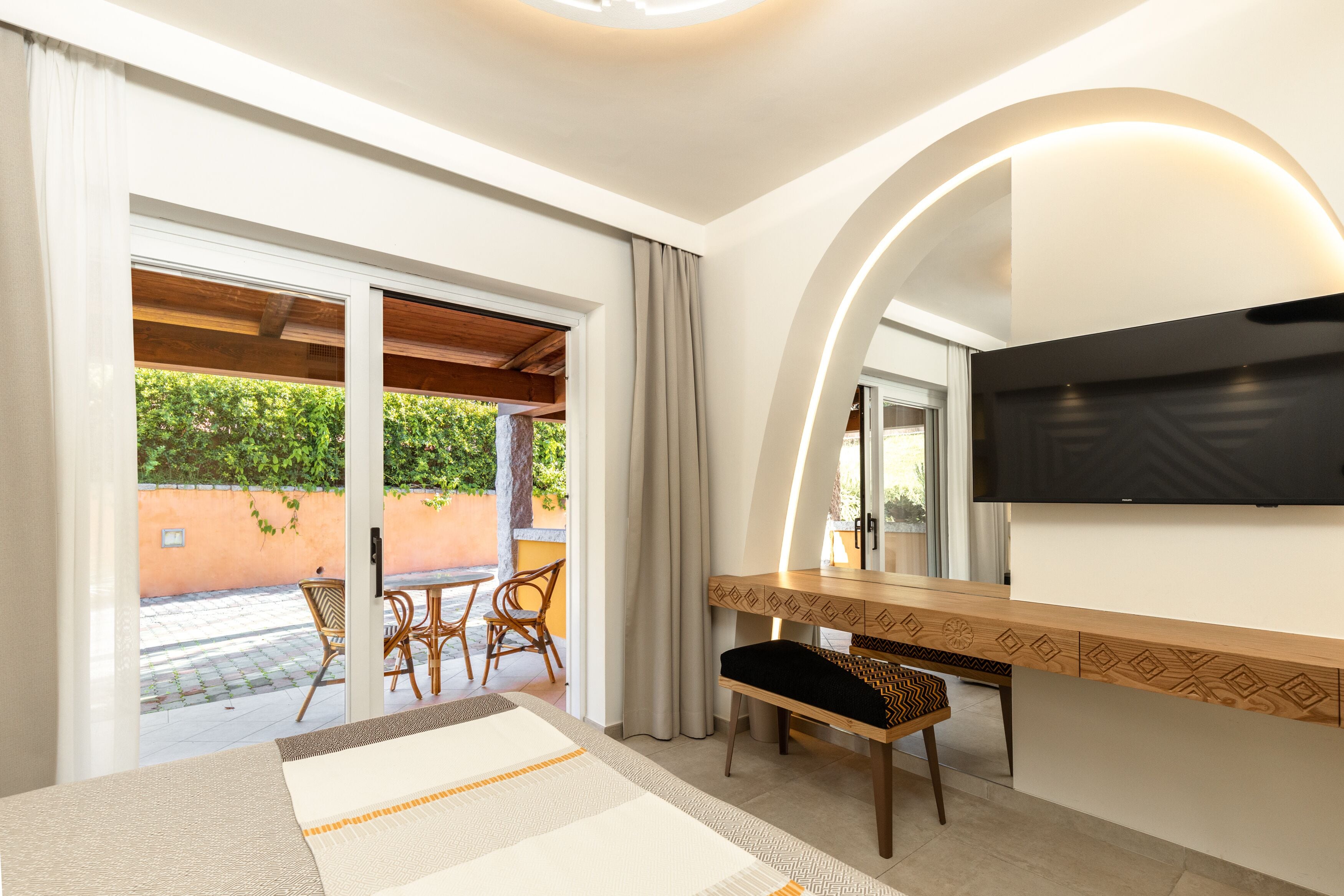 A hotel room with a large sliding glass door leading to an outdoor patio, a wooden desk with a bench, and a flat-screen TV mounted on the wall.