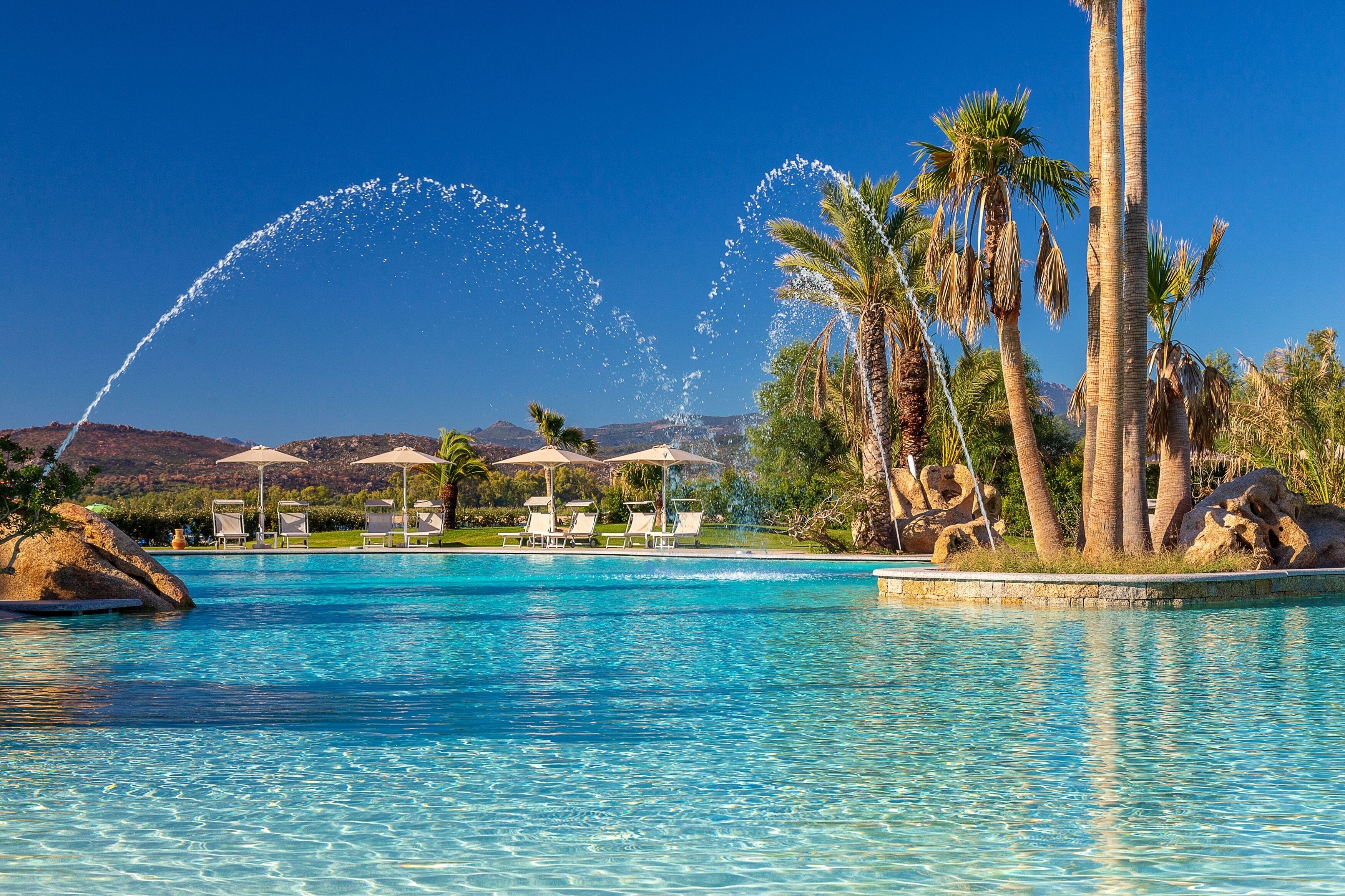 A clear blue pool with water fountains, palm trees, lounge chairs, and umbrellas.