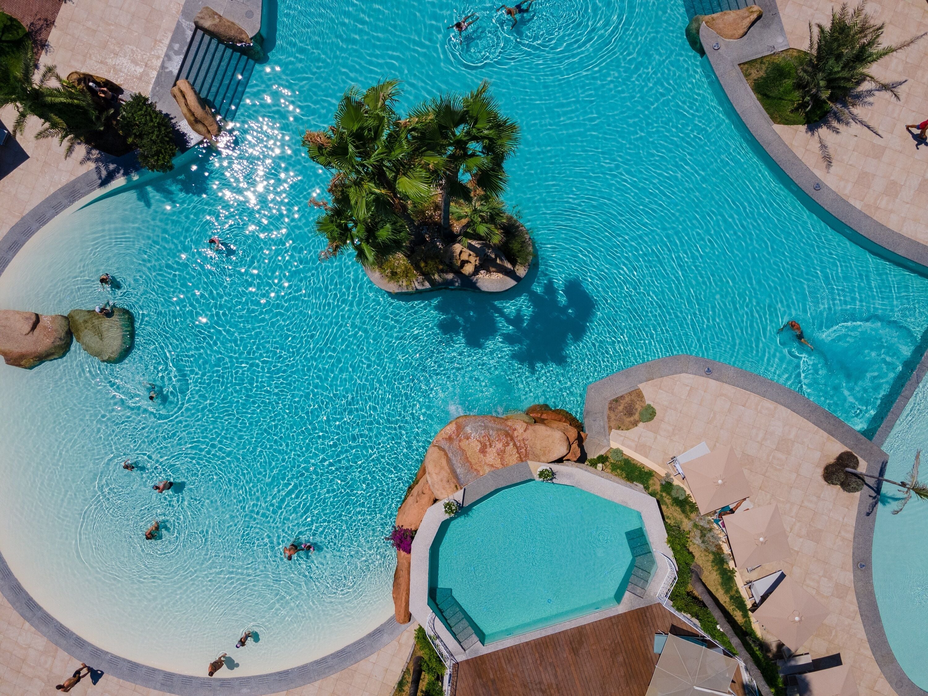 An aerial view of a large, clear blue swimming pool with multiple people swimming and lounging. There are palm trees and rocks around the pool, and a small island with a gazebo.