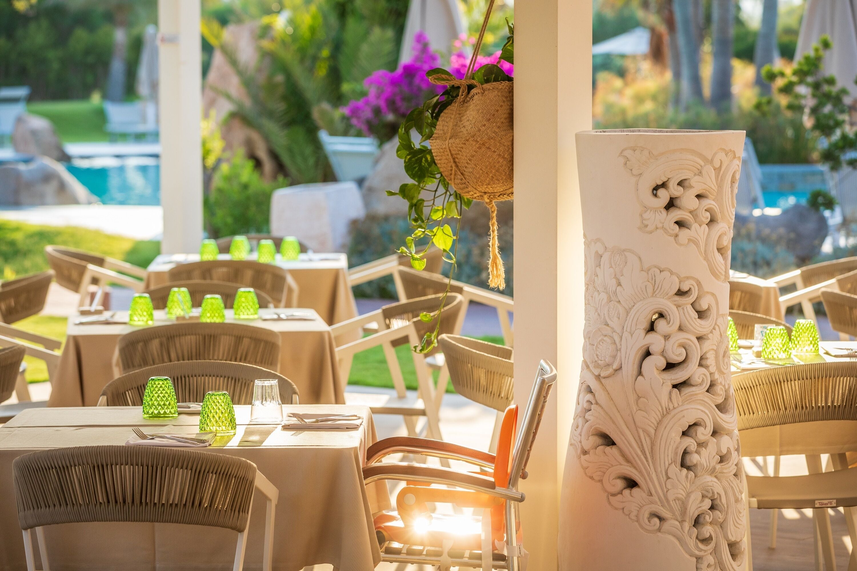 An outdoor dining area with tables set for two, surrounded by wicker chairs, and a decorative column with intricate carvings.