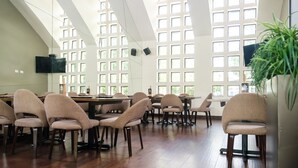 Une pièce spacieuse avec de hauts plafonds et de grandes fenêtres, un plancher en bois, ainsi que plusieurs tables et chaises disposées pour les repas ou les réunions.