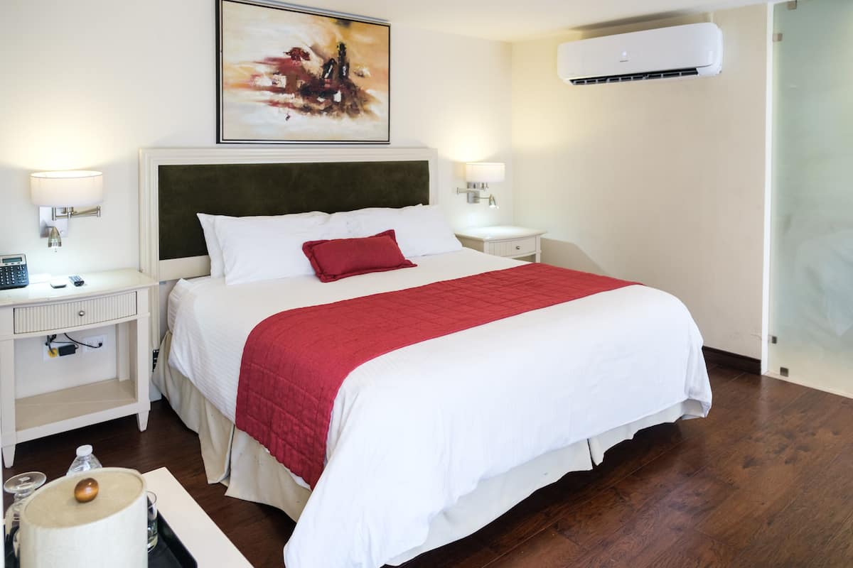 Une chambre d’hôtel avec un grand lit, du linge de lit blanc, un plaid rouge, des lampes de chevet, une table de chevet, un climatiseur fixé au mur et un tableau au-dessus du lit.