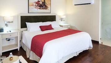 Habitación de hotel con una cama grande, ropa blanca, una manta roja, lámparas de noche, una mesita de noche, aire acondicionado de pared y un cuadro sobre la cama.