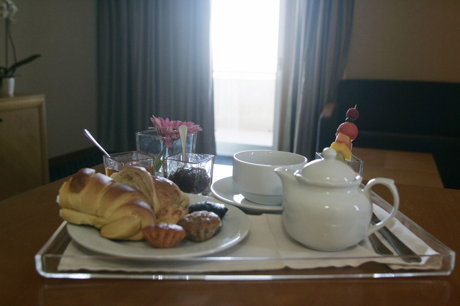 A breakfast tray with croissants, pastries, a teapot, cups, and a flower vase.