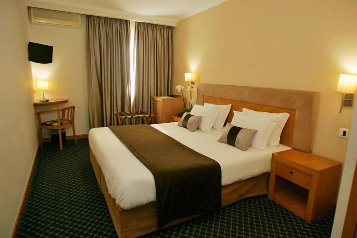 A hotel room with a large bed, a desk, a chair, a TV, and a window with curtains.