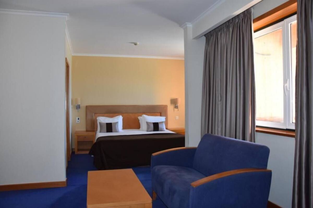 A hotel room with a bed, a blue armchair, a wooden nightstand, and a window with curtains.