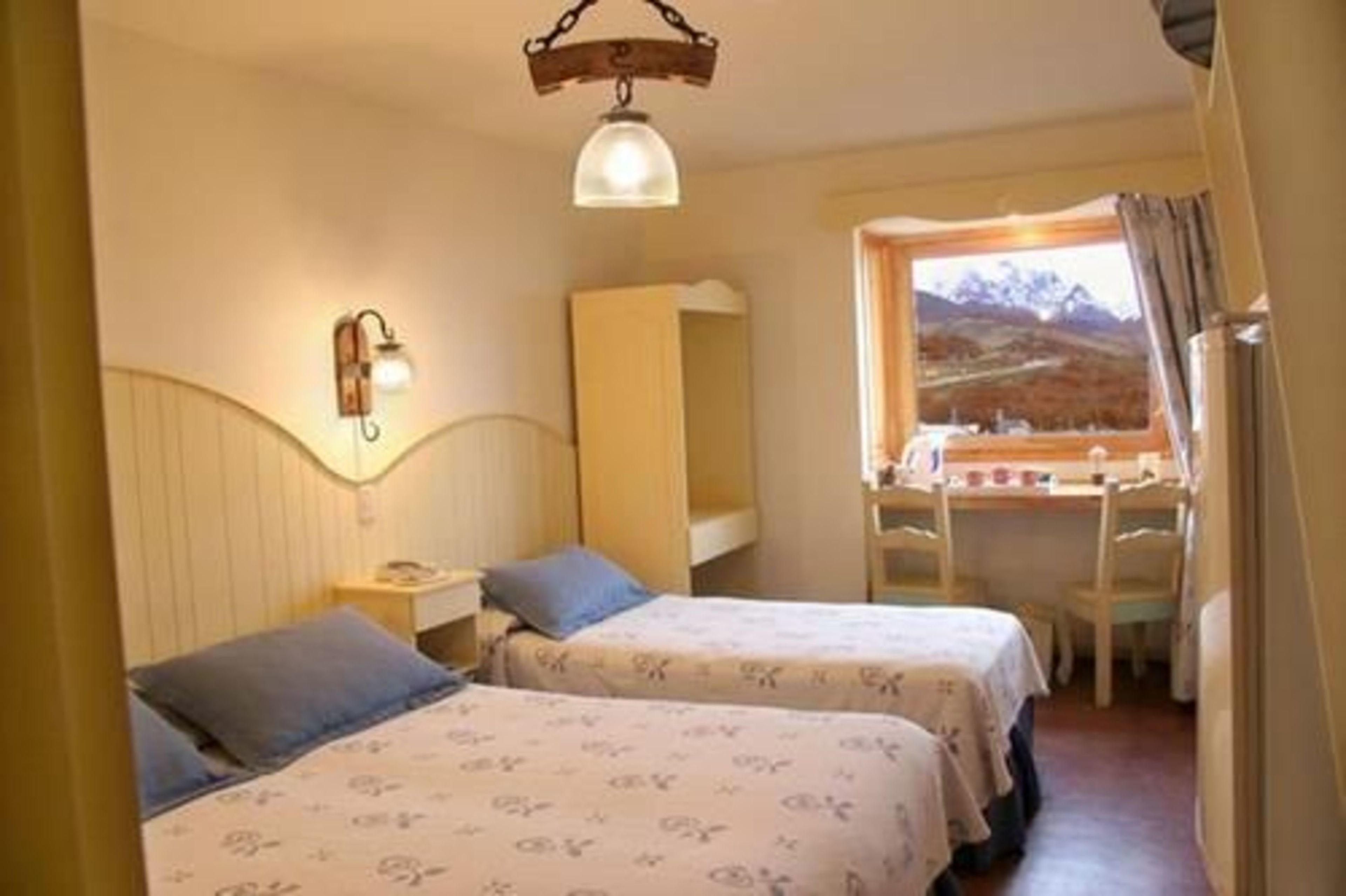 A room with two beds, a wooden desk, and a window with a view of a mountainous landscape.
