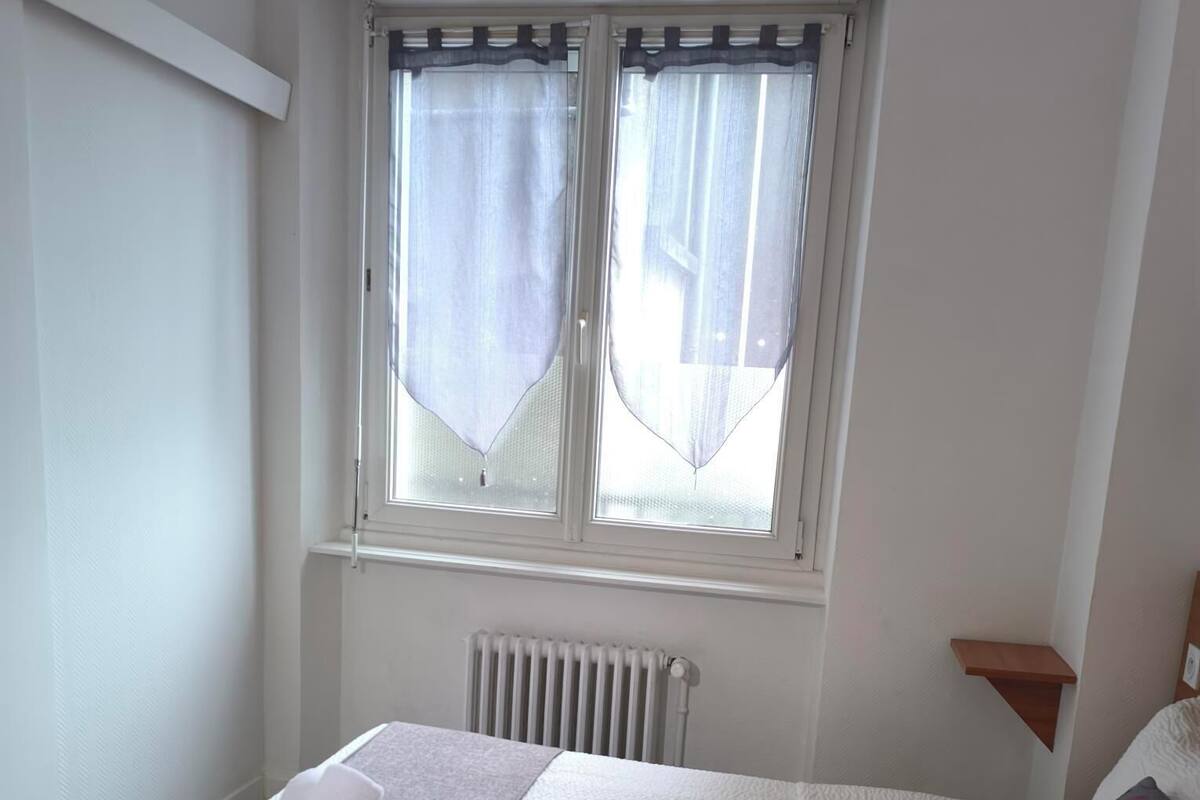 A neatly made bed with a grey and white duvet, a small bedside table with a lamp, and a window with sheer curtains.