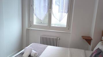 Un lit bien fait, avec une couette grise et blanche, une petite table de chevet avec une lampe, et une fenêtre ornée de rideaux légers.