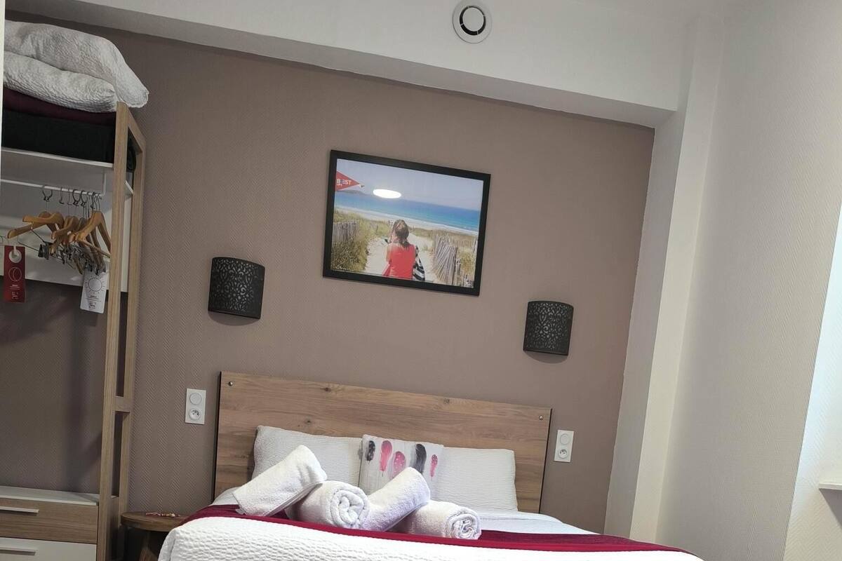 A neatly made bed with white and red linens, a wooden headboard, and a wall-mounted TV displaying a beach scene.
