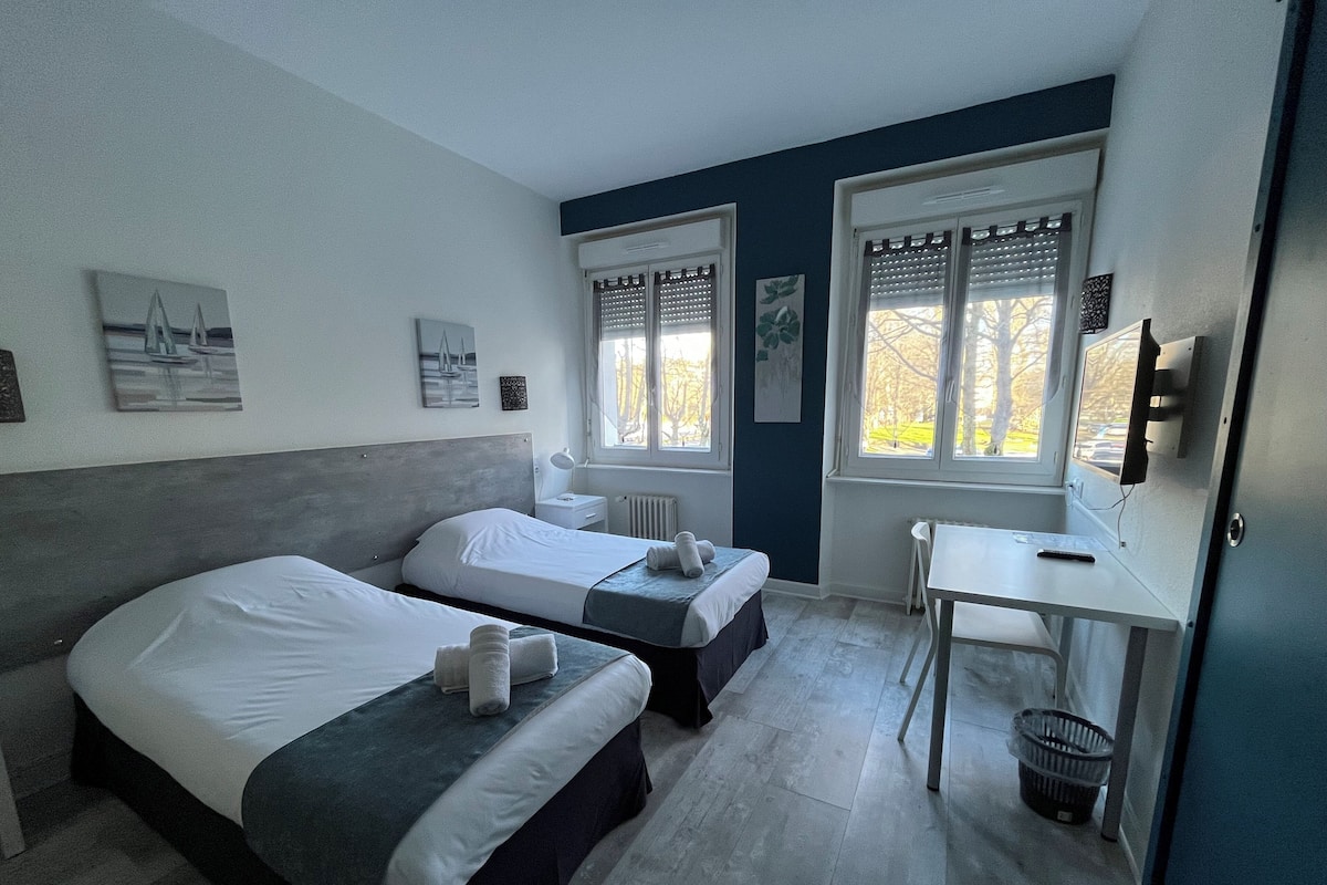 A hotel room with two single beds, a desk, and a chair. There are two windows with blinds, a radiator, and a television on the wall.